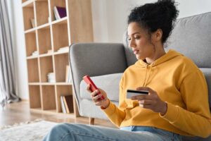 A person reviewing a checking account balance on a smartphone while sorting household bills at a kitchen table.