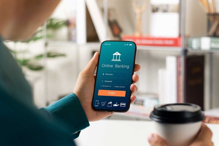 A person holds a smartphone displaying a banking app, with a home desk and documents visible in the background.